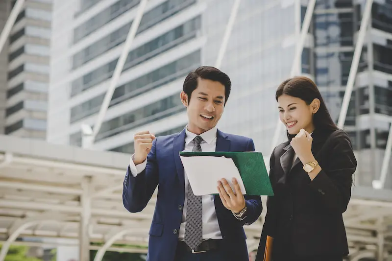 Leoker professional business team celebrating success outside modern corporate building, reviewing positive business results with enthusiasm while holding documents in a dynamic urban setting